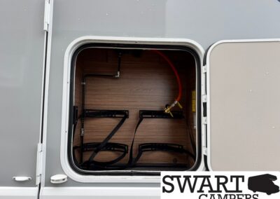 Storage hatch of a camper with interior wood-panel walls, black strap racks, and an orange hose connected to a brass fitting inside the compartment.