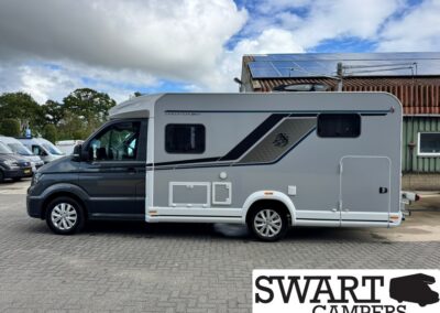 Side view of a silver-gray motorhome with black decorative stripes parked on a paved lot, with a building and blue sky in the background.