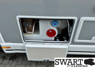 Outdoor service hatch of a camper van showing blue water-fill cap and red valve handles inside the compartment.