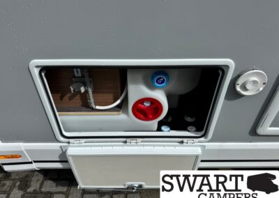 Open exterior service hatch on a camper trailer showing a blue water-fill cap and a red port with hoses inside.