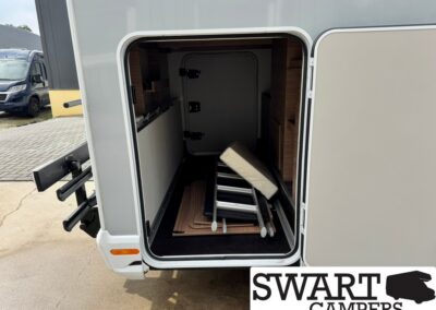 Open storage bay of a motorhome with a folded ladder resting on the floor and wooden shelving inside the compartment