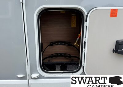Open camper storage bay with wood paneling, two black curved rack bars, and a safety valve on the right.