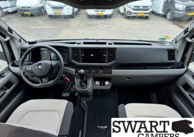 Interior view of a camper van cockpit: steering wheel, center touchscreen, climate controls, gear lever, and two gray cloth seats with a parking lot visible through the windshield.