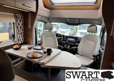 Interior of a campervan dining area with a table set for two: plates with croissants, mug, glass, and a fruit bowl; front seats visible with a skylight above.