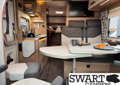 Cozy camper van interior with a small kitchen, dining table set for two, and a bed area in the back; warm wood cabinetry and seating create a homey space.