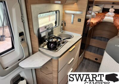 Compact camper van kitchenette with a kettle on the stovetop, stainless sink, wood cabinets, and a small counter; bed area with orange bedding and teddy bears in the background.
