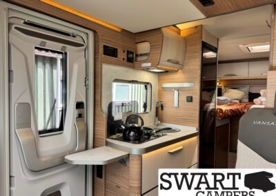 Interior of a compact camper van showing a wood-tone kitchenette with a sink, two-burner stove, and a black kettle on the stove; folding table extended from the counter and a door to the left. In the background, a sleeping area with a bed and pillows is visible.