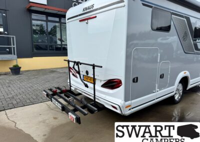 White Knaus camper van with a mounted rear bike rack and EU license plate parked beside a building, Swart Campers logo in corner.