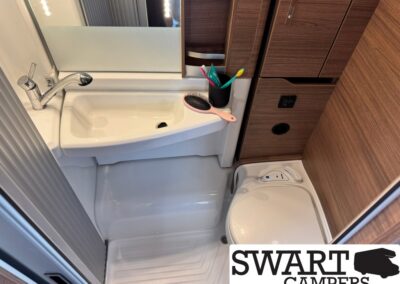 Compact camper bathroom with a white sink, chrome faucet, and mirror; toothbrush cup with toothbrushes and a pink hairbrush on the counter, wooden cabinets nearby, and a small toilet to the right; SWART CAMPERS logo in the bottom corner.