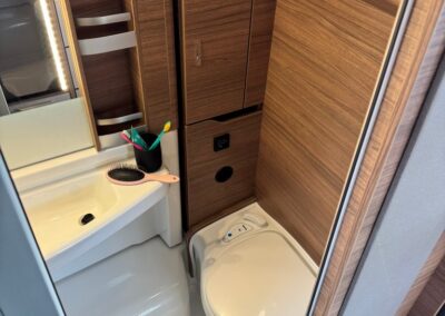 Compact camper bathroom with woodgrain cabinets, a small sink, and a closed toilet, toothbrushes resting in a holder on the counter.