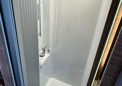 Compact white shower enclosure in a camper/RV with a chrome faucet and vertical grab bar under overhead lights
