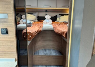 Interior of a camper van bedroom with two twin bunks, orange polka-dot bedding, and teddy bears on pillows under overhead cabinets. A small skylight above and wood paneling give a cozy, compact feel.