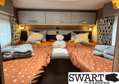 Cozy camper bed setup with orange polka-dotted bedding, gray pillows, and a teddy bear at the head of the bed.