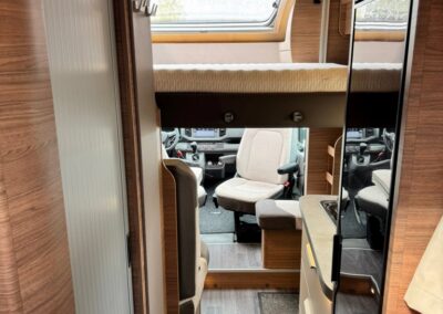 Interior of a camper van showing a raised bed over the cab, wood paneling, and a compact kitchenette along the right wall; driver seats visible in the cab area.