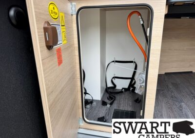 Open camper storage cabinet with an orange hose, black cables, and securing straps inside a small compartment.