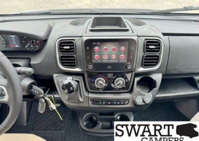 Front interior of a camper van dashboard with a central touchscreen display, climate controls, and gear shifter visible between the seats.