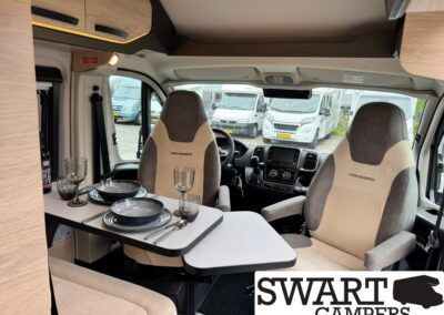 Camper van interior with a table set for two: plates, glasses, and cutlery; front seats and cockpit visible; SWART CAMPERS logo in corner.