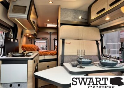Cozy camper van interior with a kitchenette on the left, a rear bed with orange polka-dot bedding, and a dining table set with blue dishes and glassware.