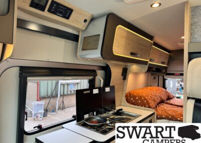 Camper van interior with a compact kitchen (two-burner stove) and overhead cabinets, plus a bed with orange polka-dot bedding nearby.