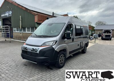 Silver camper van parked in a lot near a garage building with SWART CAMPERS logo in the corner.