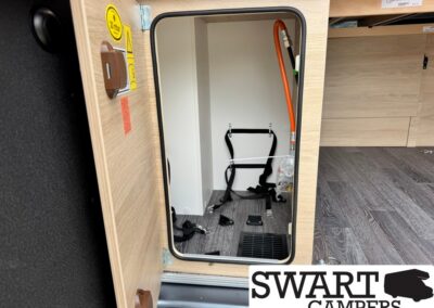 Open storage cabinet in a camper interior, showing black strap restraints on the back wall, an orange gas hose on the right, small parts on the floor, and a floor vent.