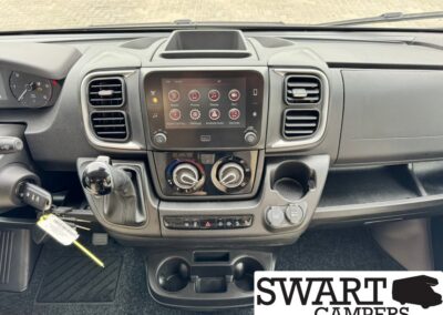 Front cabin of a van showing a center infotainment touchscreen, climate control dials, gear shifter, cup holders, and ignition keys; SWART CAMPERS watermark in the bottom right.