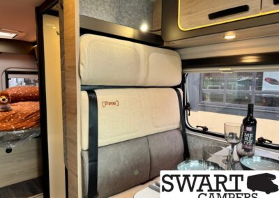 Interior of a camper van: cream cushioned bench seating, a small table with wine glass and bottle, and a bedroom in the back; logo SWART CAMPERS overlays the corner.