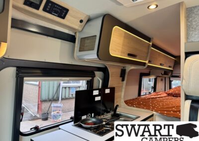 Interior of a camper van showing a compact kitchenette with a stove and sink, upper storage cabinets, and a sleeping area with an orange polka-dot bedspread.