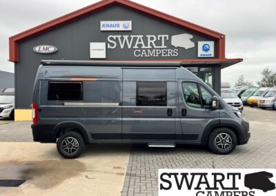 Gray camper van parked in front of a Swart Campers building with a red gabled roof and large white SWART CAMPERS sign above the entrance, side windows visible.