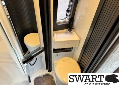 Compact camper bathroom with a round toilet beside a small sink and a window, flanked by dark vertical panels.