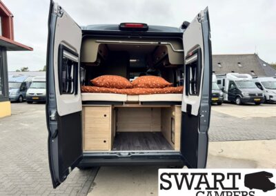 Camper van rear with doors open, showing a bed with orange polka-dot pillows and wooden storage cabinets underneath.