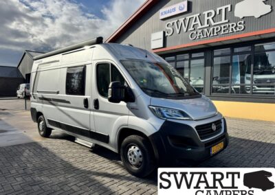 Silver camper van parked outside a Swart Campers showroom, with a large Swart Campers sign on the building nearby.