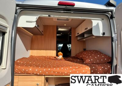 Camper van interior with a brown polka-dot bed, wooden cabinets, and a small plush toy on the mattress; SWART CAMPERS logo in corner.