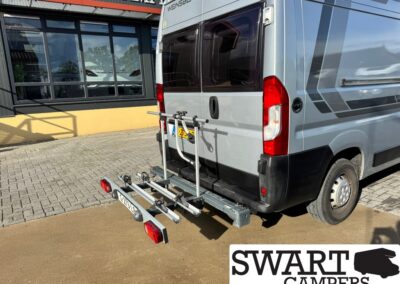 Rear view of a gray camper van with a two-bike rack mounted on the hitch, parked outside a camper shop; SWART CAMPERS logo visible in the bottom-right corner.
