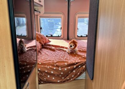 Interior of a campervan bedroom with a brown polka-dot bed, plush lion toy, and wood cabinetry along the sides; windows at the back let in light.