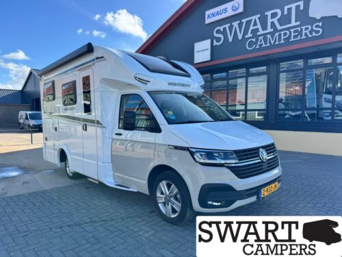 White VW camper van with an over-cab sleeper parked in front of a Swart Campers showroom, with Knaus branding visible on the building.