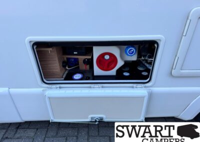Exterior service hatch of a motorhome opened to reveal water tank fill caps, valves, and storage shelves inside the bay.