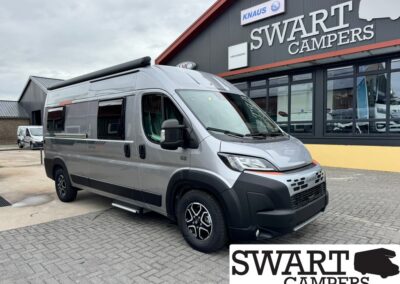 Silver camper van parked in front of a SWART CAMPERS storefront, showing a black bumper and side steps under a cloudy sky.