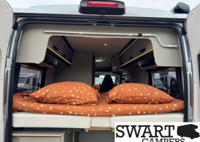 Camper van rear bed with orange polka dot bedding and overhead storage cabinets.