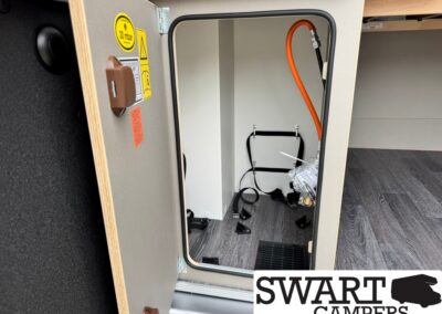 Open camper storage cabinet with an orange hose, electrical cables, and mounting hardware visible inside. Swart Campers watermark appears in the corner.