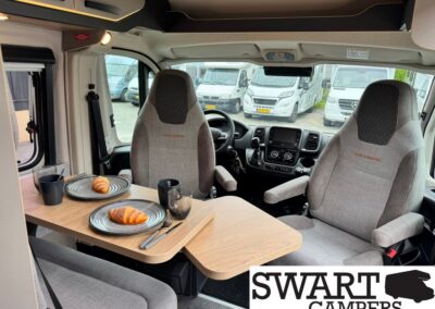 Inside a camper van, a wooden dining table set for two with croissants on plates and coffee mugs, facing the cockpit seats.