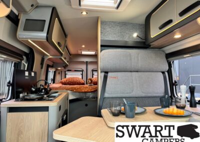 Camper van interior: kitchenette with stove, bed at rear with orange bedding, and a wooden table set for two.