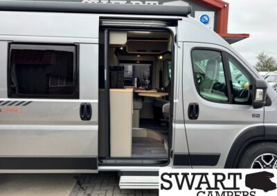 Grey camper van with side door open, showing a compact kitchen and seating area inside; parked outdoors at a dealership.