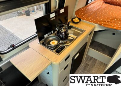 Camper van kitchenette with a two-burner stove, kettle, egg frying in a pan, a fold-out counter, and a bed with an orange polka-dot duvet nearby.