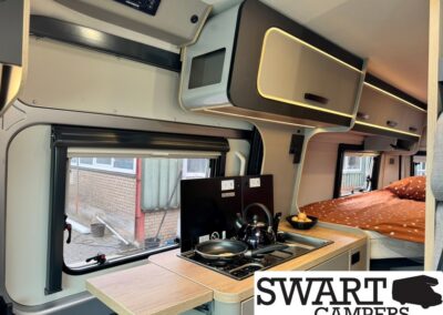 Interior of a compact camper van with a kitchen area: stove, kettle, frying pan, and fold-out counter beside a bed with orange bedding.