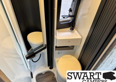 Compact camper bathroom: toilet beside a small sink with a black faucet, window above, and dark paneling on the walls.