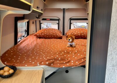 Compact camper bedroom with orange polka-dot bedding and a small plush lion on the bed; kitchenette and eggs in a basket visible nearby.