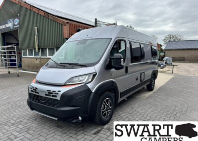 Silver camper van parked on a paved lot beside an industrial building, showing a high roof and side windows (Swart Campers).