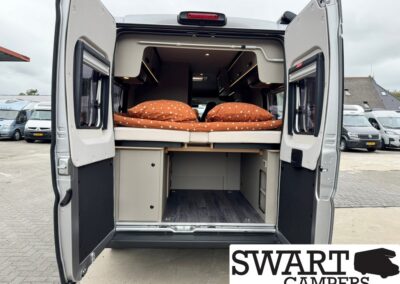Rear of a campervan with the back doors open, showing a raised bed with orange bedding and built-in storage underneath.