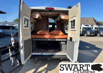Rear of a white camper van: bed with orange polka-dot bedding, wood cabinets, and a bike rack on the left, SWART CAMPERS logo bottom right
